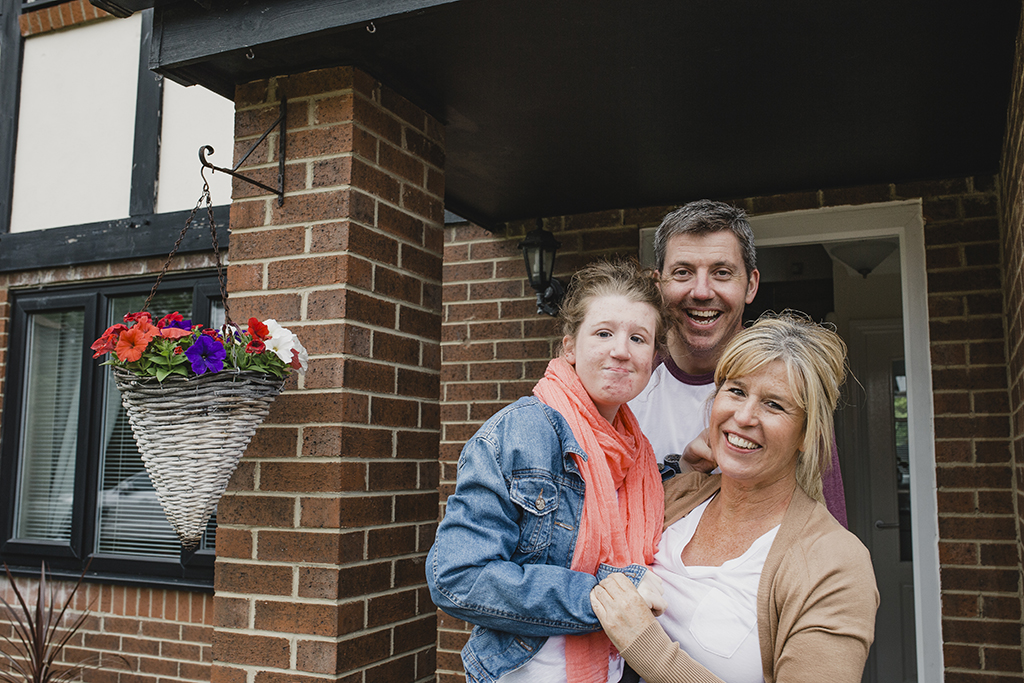 Portrait of a family of three outside their home. The child has cerebral palsy and is being held by her mother.