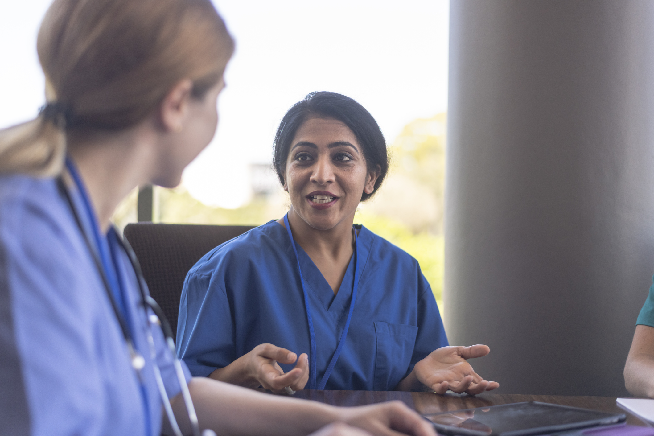 Two nurses in blue scrubs talk at conference table