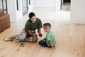 The mid adult female soldier sits on the wood floor of the living room and helps her elementary age son figure out how to put some toy cars together.
