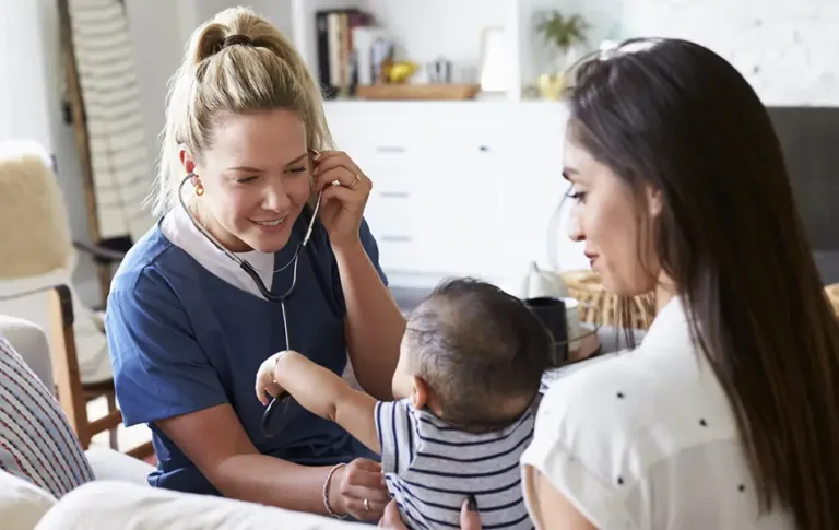 Nurse using a stethoscope to engage with a baby while speaking to a smiling mother at home — value-based pediatric healthcare solutions from Maxim Healthcare.