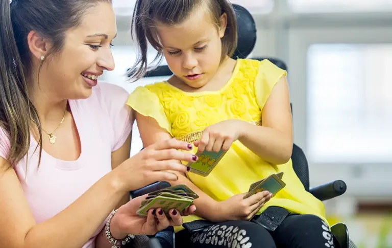 Happy caregiver engaging in a fun card game with a young girl in a wheelchair, showcasing the supportive and nurturing environment provided by Maxim Healthcare caregivers.