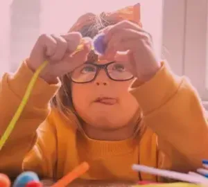 Young girl wearing glasses and a yellow sweater plays with colorful craft materials, focusing intently.