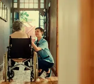Caregiver crouching to speak with an elderly woman in a wheelchair in a warmly lit hallway.