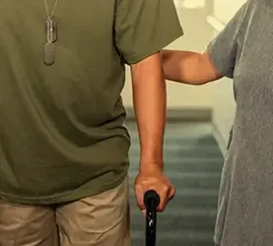 veteran using a cane being supported by a companion while walking down a hallway