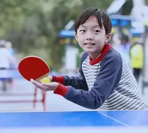 Young boy smiling while playing table tennis outdoors, holding a red paddle and yellow ball.