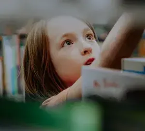 Young girl with long brown hair looking up attentively in a classroom or library setting, with bookshelves in the background.