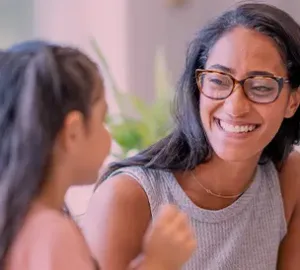 Smiling woman with glasses interacting with a young girl in a bright, friendly indoor environment.