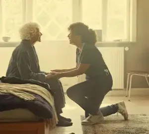 Caregiver kneeling beside an elderly woman, holding hands in a warmly lit bedroom setting.