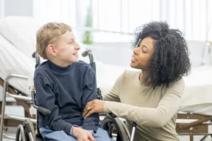 Young boy sitting in his wheelchair smiling while looking at a smiling woman next to him.