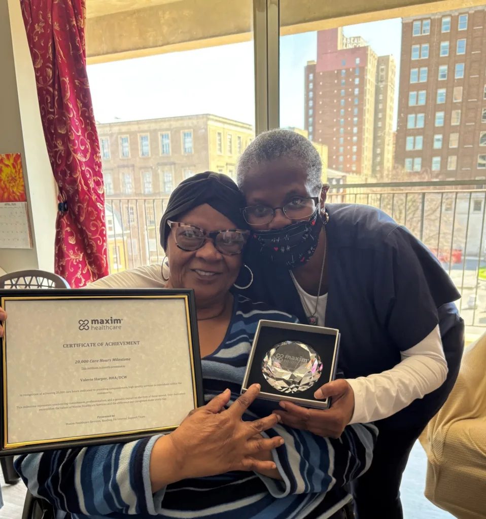 Caregiver holding a Maxim Healthcare 20,000 Care Hours Milestone certificate and crystal award while posing with her patient in front of large windows overlooking a city street.